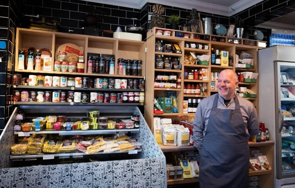 Larry at the grocery shelves in Market Fair, Marino — cheeses, jars and dry goods behind the counter